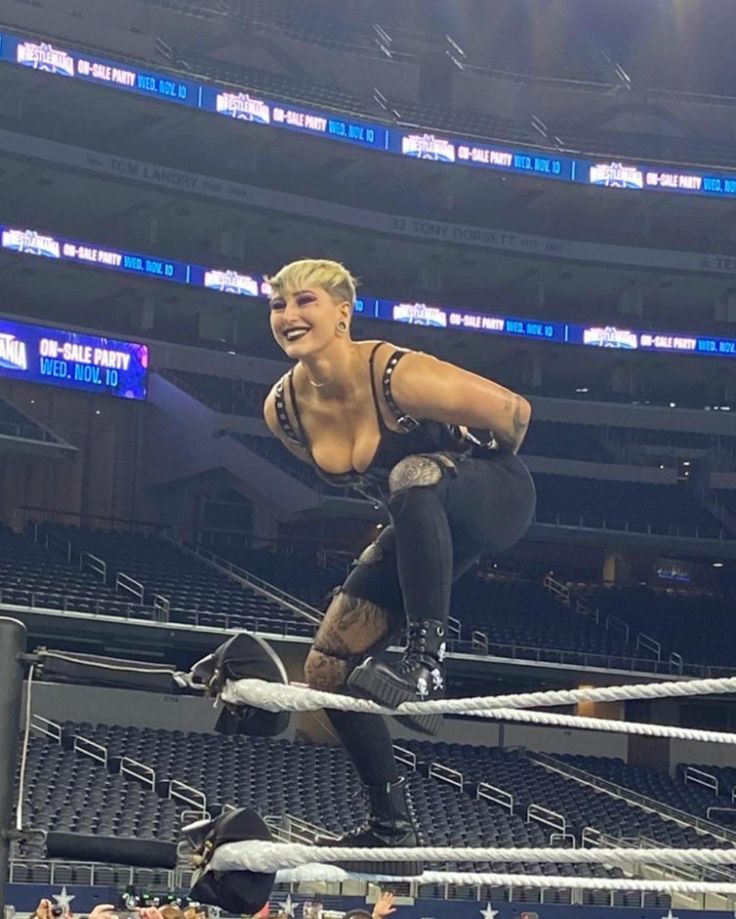 a female wrestler in a black outfit is poised on the top rope of a wrestling ring, looking down at the crowd. the arena is filled with people, and the wrestler is in a powerful pose, ready for action.