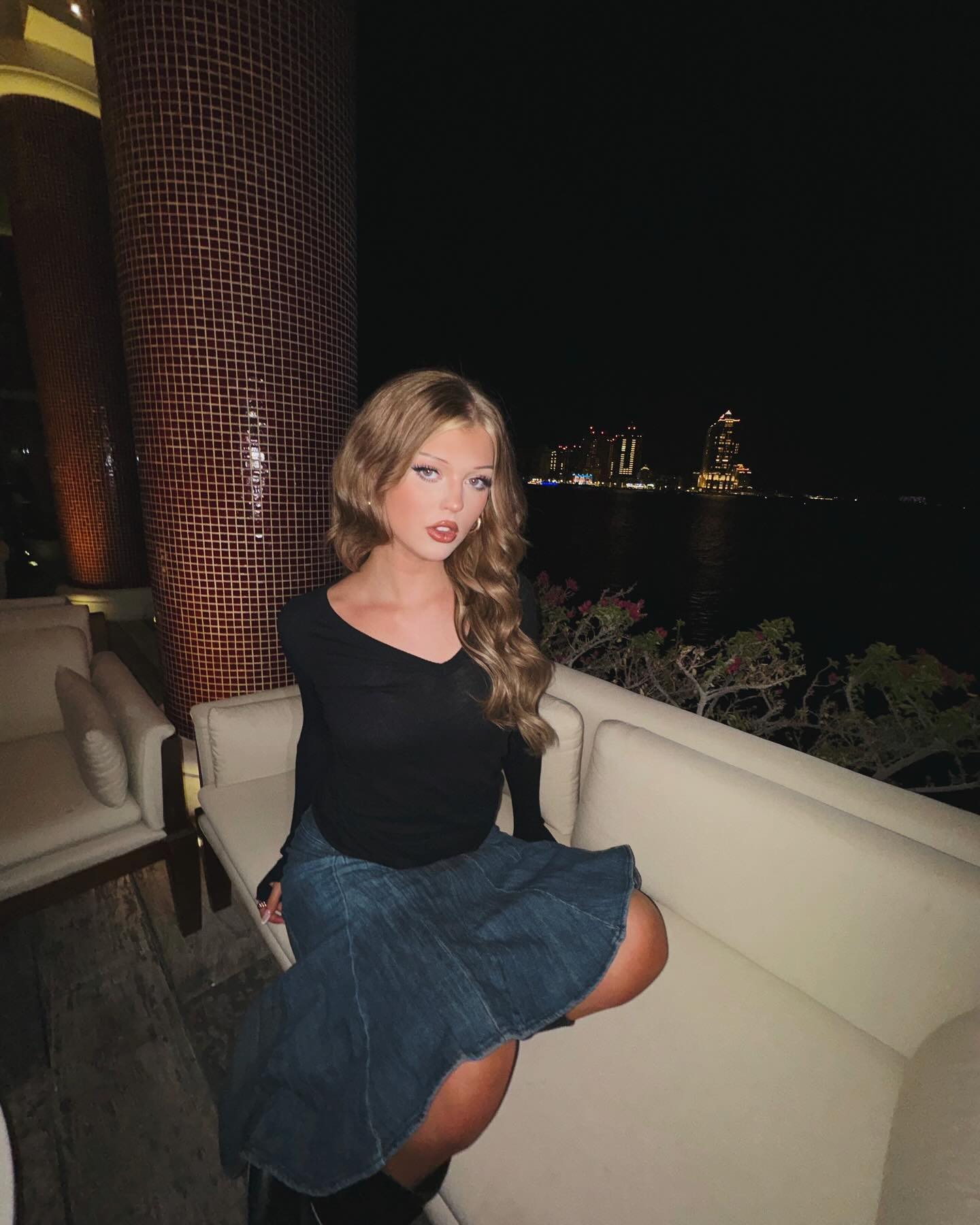 a young woman with blonde hair sits on a white couch outdoors at night. she is wearing a long sleeve black shirt and a denim skirt. in the background, there is a city skyline.