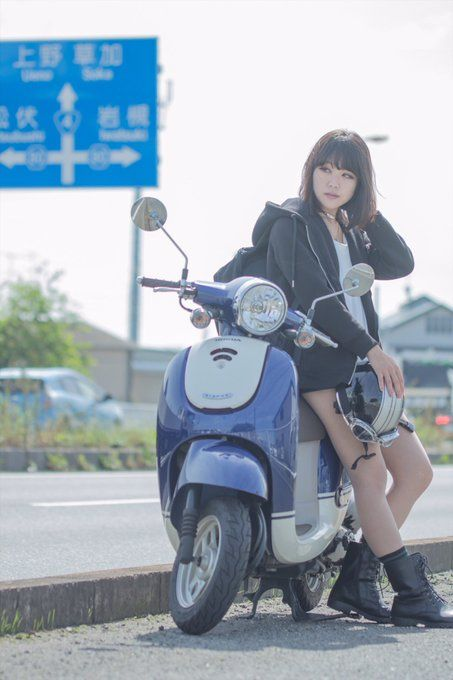 a young woman with short, dark hair stands next to a blue and white scooter parked on the side of a road. she is wearing a black hoodie, a black skirt, and black boots. she is holding a white helmet in her right hand and is looking at the camera. the background includes a blue road sign, a street with cars, and buildings. the photo is taken during the day with natural lighting.