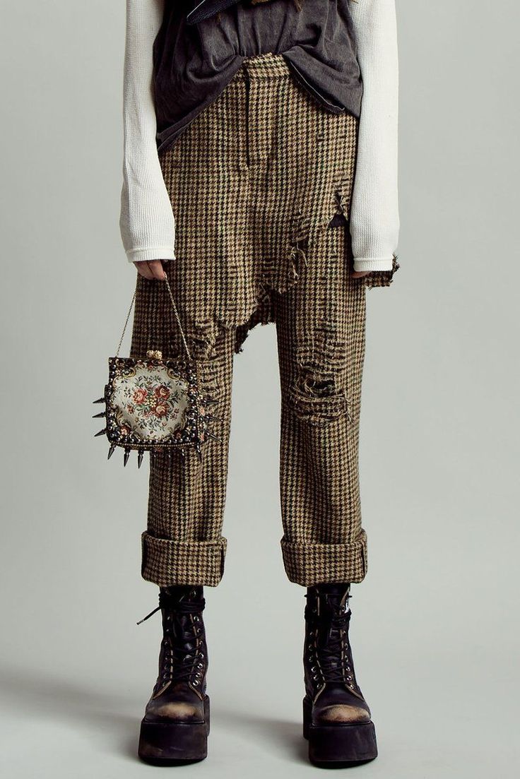 a person wearing a white longsleeved top, a dark vest, distressed plaid trousers, and black platform boots stands in front of a light gray background. they are holding a small square handbag with a floral pattern and studded edges. the trousers are cuffed at the ankles and have several distressed areas with holes. the overall style is grunge and edgy.