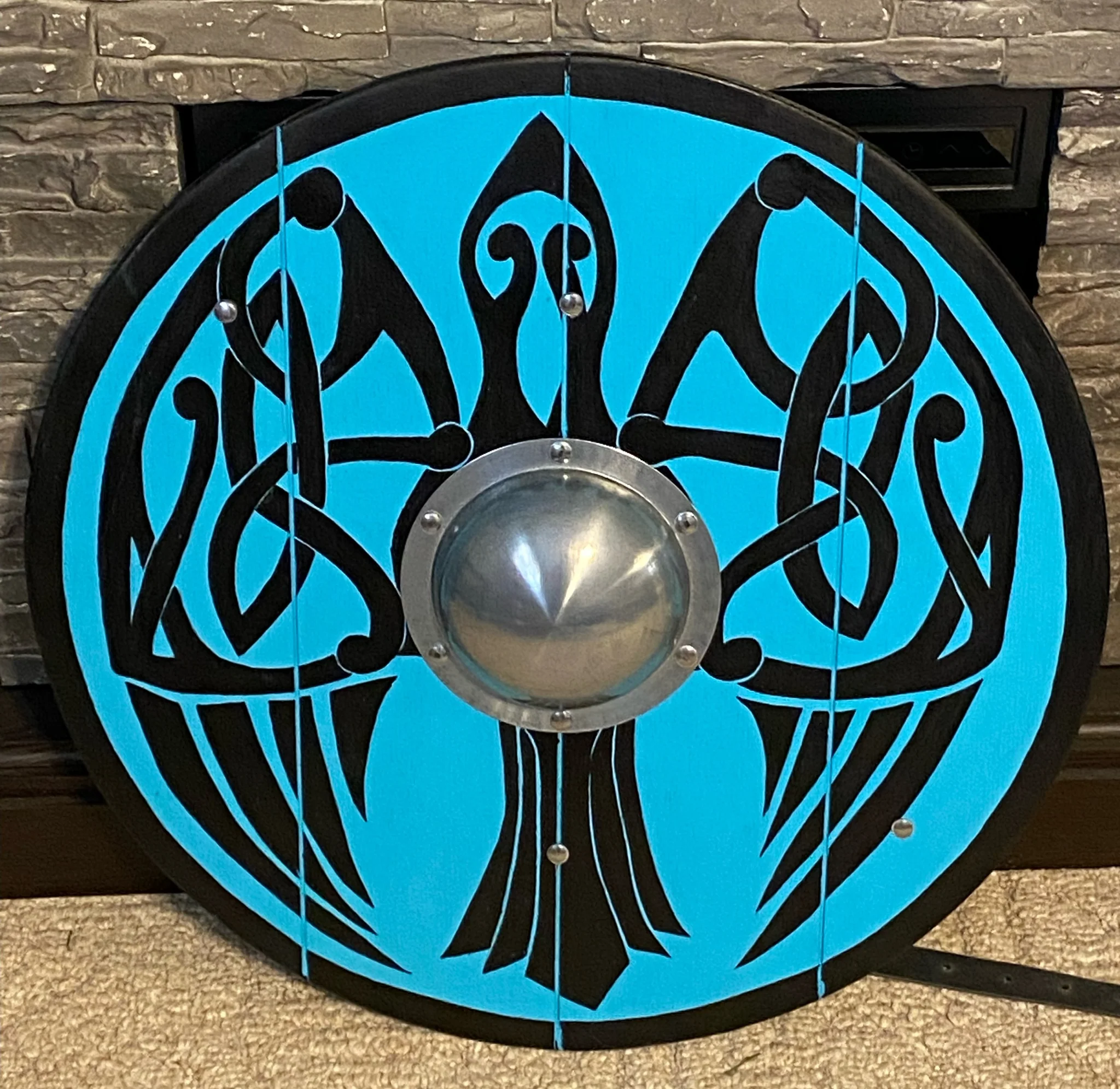 a round viking shield painted with a stylized black raven on a light blue background. the raven's wings are decorated with celtic knotwork patterns. the shield has a central metal boss and wood grain showing on the edges.