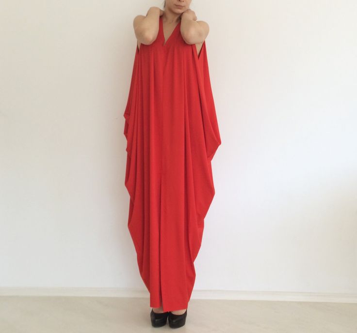 a woman is standing in front of a white wall, wearing a long, loosefitting red dress. she has her hands up near her neck and is wearing black shoes. the style is simple and casual.