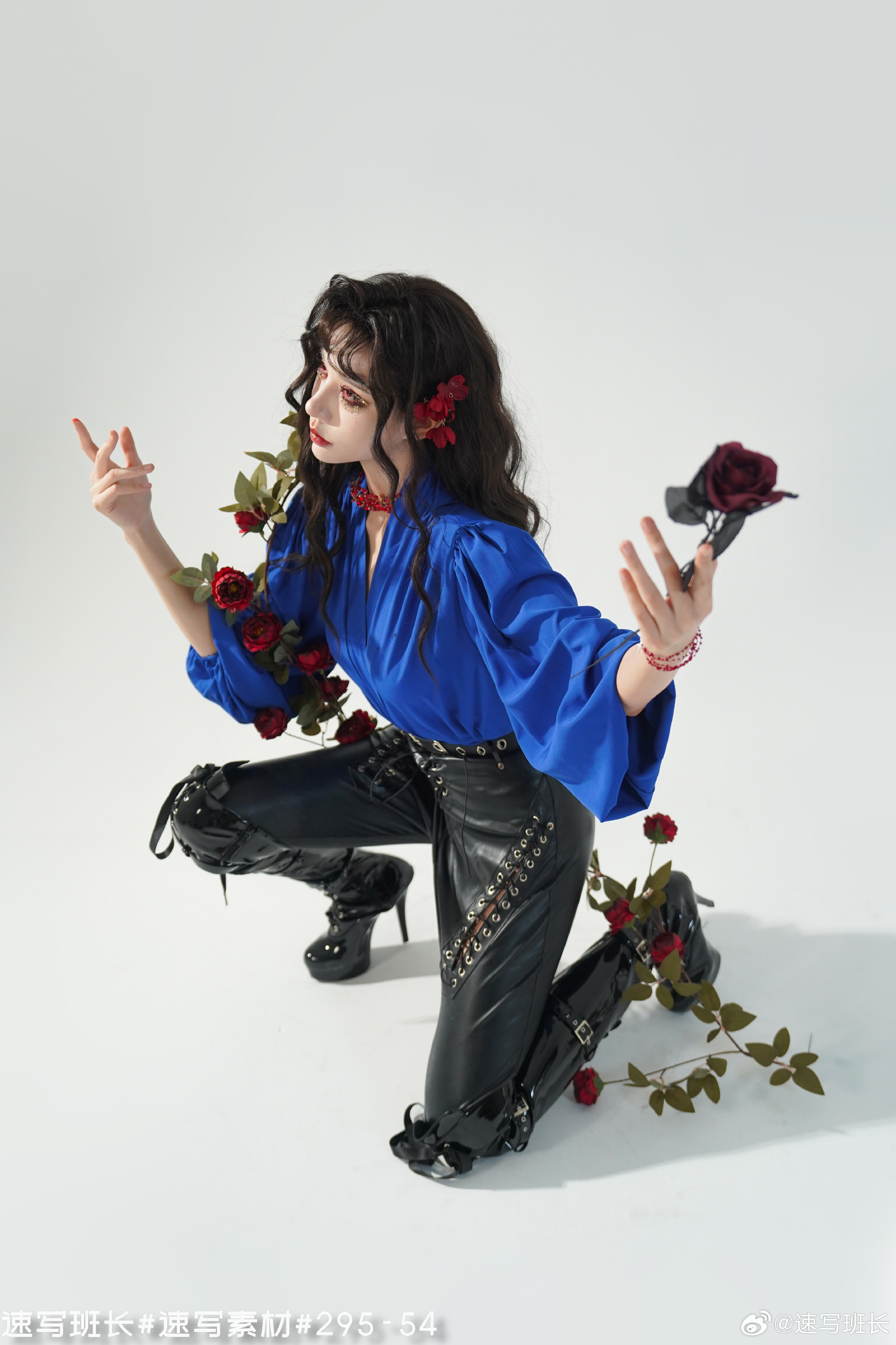 a woman in a blue blouse and black leather pants strikes a dramatic pose against a white background. she is adorned with red roses that are wrapped around her arms and legs. her dark, curly hair is styled with two red roses. the lighting is artificial, and the image is taken from a high angle.