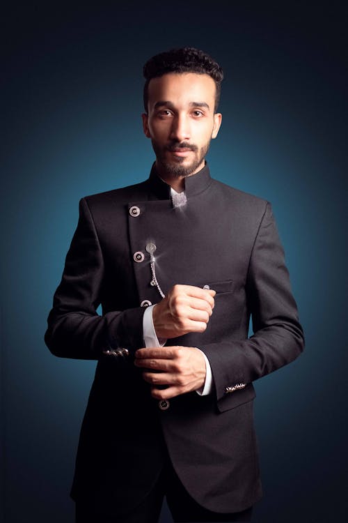 a closeup portrait of a stylish, cleancut man of color. he's dressed in a sharp, black indowestern style bandhgala suit, and is adjusting his cuffs. his expression is confident and sophisticated, emphasizing his wellgroomed appearance. the dark background enhances the overall mood of elegance. the image is welllit, with a focus on the man's features and attire, conveying a sense of professionalism and style.