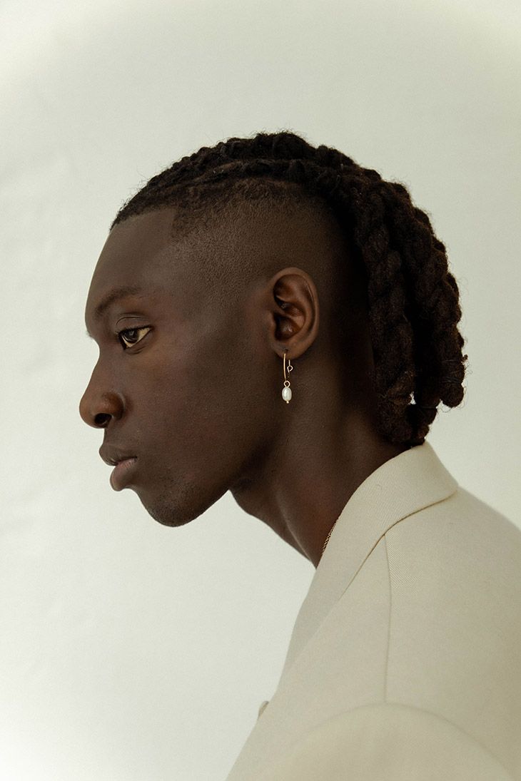 this is a studio shot of a man in a beige suit. he is shown in profile, facing left. he has short, dark hair on the sides and dreadlocks on top. the man is wearing an earring. the background is plain white.