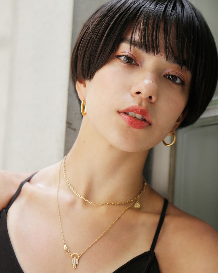 a young asian woman with short, bobbed hair with bangs smiles slightly towards the camera. she is wearing a black tank top, gold earrings, and a gold necklace. the photo is taken from her chest up and is shot at a slight angle to show the woman's left side profile. she looks to be in her late teens or early twenties. her makeup is natural and she has a radiant complexion.