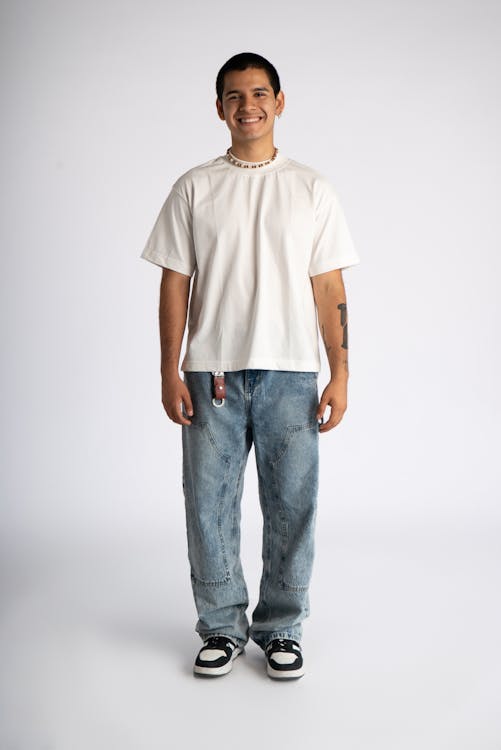 a full shot studio photograph of a young man standing and smiling in casual wear against a white background. he is wearing a white tshirt with a loose fit, light blue baggy jeans with patchwork detail, and black and white sneakers. he also has a choker necklace and a tattoo on his left arm.