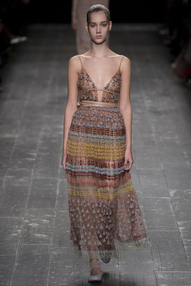 a young woman, wearing a long, brown floral lace dress, walks down a runway. she is wearing white sandals and has straight brown hair. the background is blurry and appears to be a brick floor.