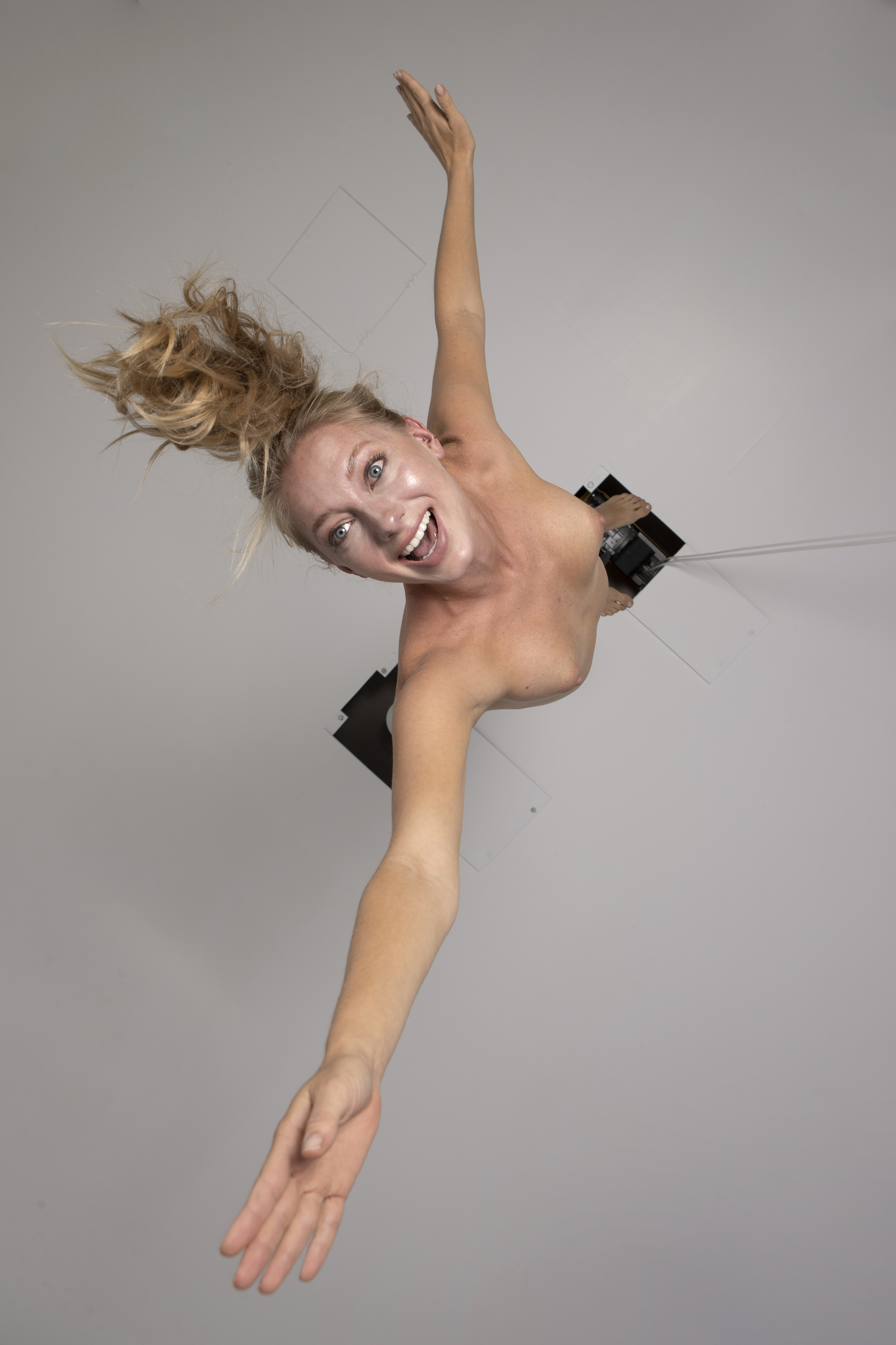 a woman with blonde hair and raised arms is smiling and looking at the camera. she is nude from the shoulders up.