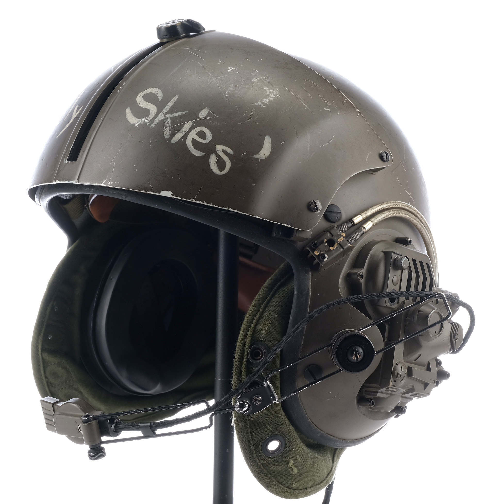 a closeup of a green military flight helmet with the word "skies" written on the side, in a worn, distressed font. the helmet is equipped with a headset and a visor, and is designed for use by pilots in the air force.
