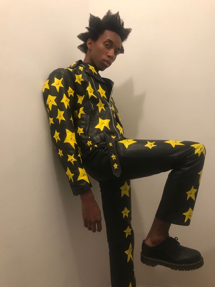 a young black man with an afro is wearing a black leather jacket and pants. the jacket and pants are decorated with a repeating pattern of yellow stars. the man is standing in a room with a white wall. he is posing with one leg bent and his foot up, looking toward the camera.
