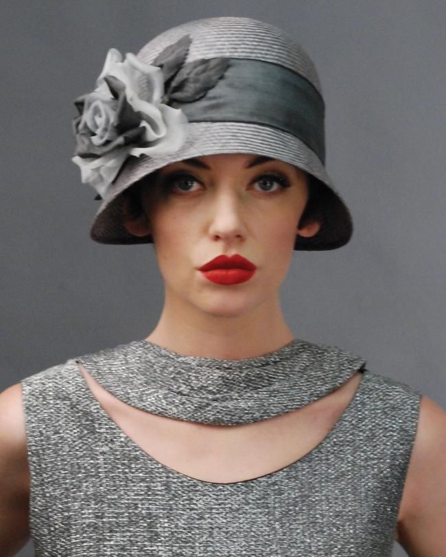 a woman in a grey cloche hat adorned with a large white rose and a grey, textured dress with a cutout detail on the neckline. the hat has a band of grey satin and her red lips are striking. she looks directly at the camera with a confident expression.