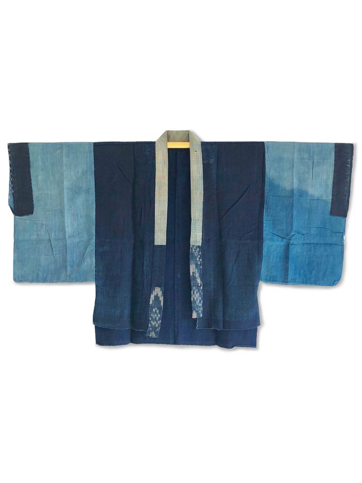 a vintage indigodyed japanese kimono or jacket is displayed flat against a white background. the garment has a traditional shape with wide sleeves and a wraparound front, featuring patchwork designs in varying shades of blue. the yoke is a lighter gray fabric, showing some traditional patterns. the garment is hung from a wooden hanger. the image highlights the handmade and artisanal aspects of the clothing.