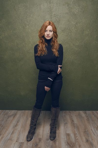 a woman with long red hair stands against a green backdrop. she is wearing a black turtleneck sweater, black jeans, and brown leather boots. she has her arms crossed in front of her and is looking slightly up and to the left. the lighting is soft and the overall tone of the image is warm.
