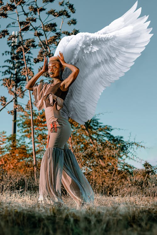 a stunning image of a darkskinned african woman adorned with large, white angel wings. she is wearing a flowing, lightbrown dress and has her hands gently placed on her head.  the setting appears to be an outdoor natural environment, a field with dry grass and trees in the background. the lighting suggests it's likely golden hour, providing a warm, ethereal feel to the photo. the overall mood is mystical and powerful, portraying the woman as a strong, graceful figure. the image is a striking example of artistic portrait photography, highlighting the beauty of the model and the symbolism of the wings.