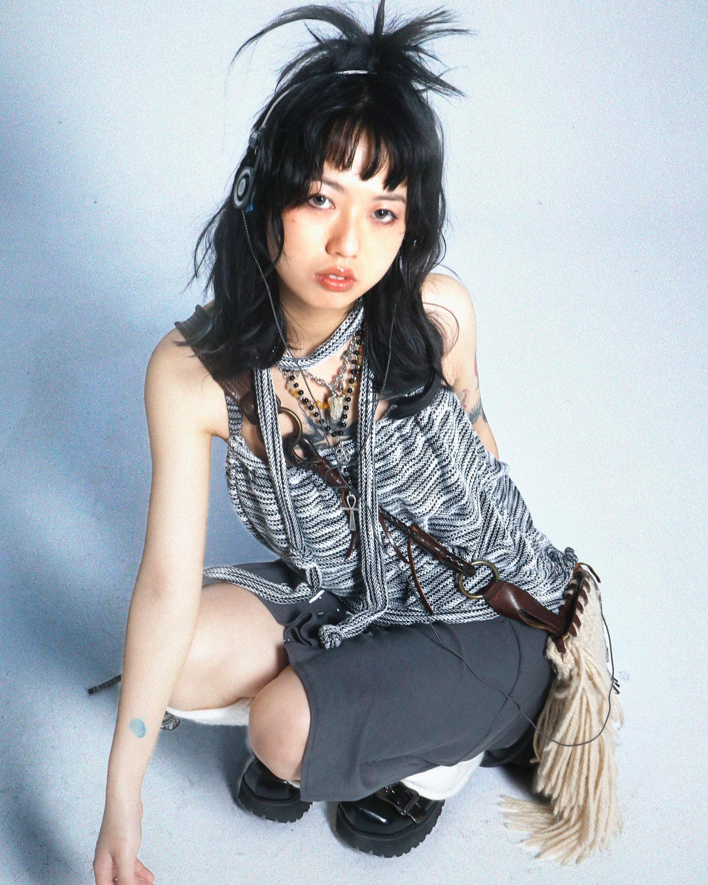 a young asian woman with a unique edgy style poses for a studio portrait. she's sitting, wearing a gray knit top, dark gray shorts, and several accessories, including headphones, necklaces, and a fringe bag. her hairstyle is spiky and dark, and her makeup is minimal but effective. the background is a plain white, allowing the model and her outfit to stand out. the overall mood is stylish and alternative, showcasing a modern and individualistic fashion sense.