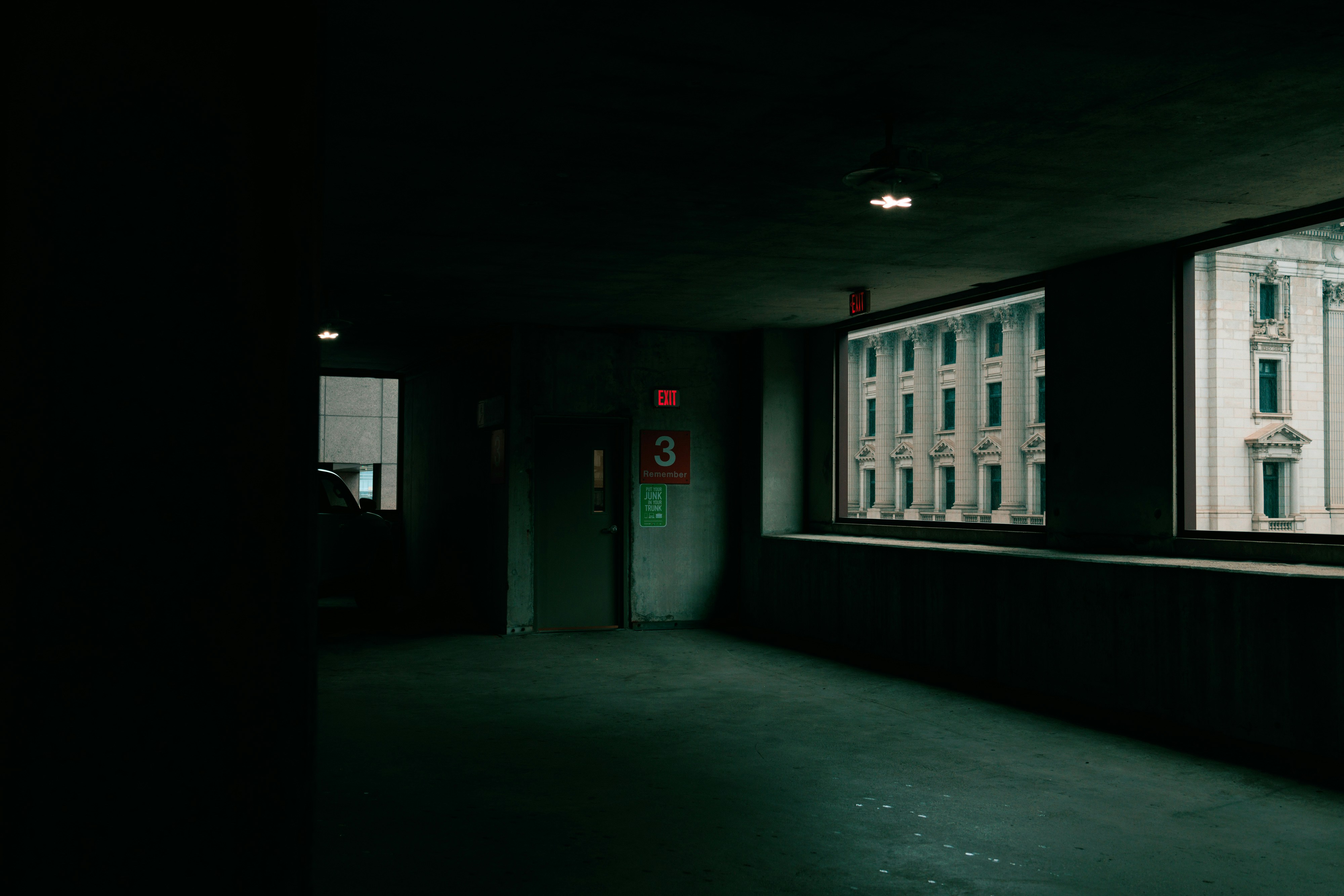 an empty parking garage shrouded in shadows, with a lone lit exit sign breaking the darkness. through the large windows, a glimpse of an ornate building facade bathed in the warm glow of the outside world contrasts with the cool, somber tones of the garage's interior.