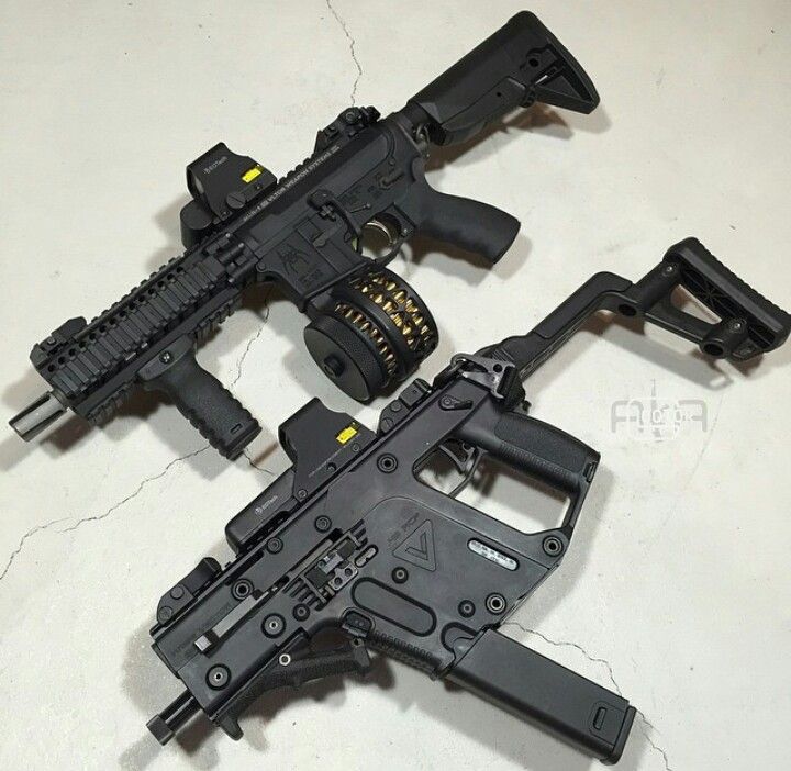 two black firearms are laying on a light gray surface. the top firearm is an ar15 style rifle with a drum magazine, optic, and foregrip. the bottom firearm is a kriss vector with an optic and a magazine.