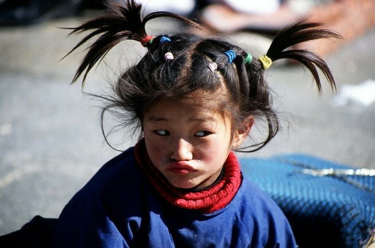 a young girl with dark hair styled in pigtails held together by colorful hair ties. she is wearing a blue shirt with a red turtleneck sweater. she is looking to the side with an expression that appears to be annoyed. she is outside and the background is out of focus.