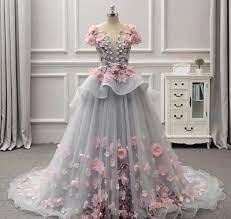 the image showcases a stunning, floorlength gown on a dress mannequin. the dress is predominantly light gray tulle, adorned with lavish 3d pink and light gray floral appliques that cascade down the skirt and bodice.  it features an offtheshoulder neckline with a sweetheart bodice and a layered, ruffled skirt. the dress is displayed in an elegant interior setting with a white cabinet visible in the background. the overall style evokes a romantic, fairytale aesthetic suitable for a wedding, prom, or other formal occasion.