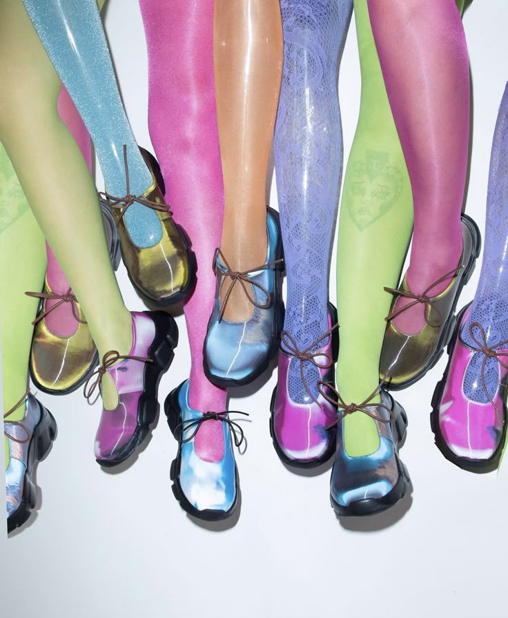 a group of legs wearing colorful glossy tights and chunky soled mary jane shoes with tieup closures. the tights and shoes are in various metallic and colorblocked combinations, creating a vibrant and fashionable display.