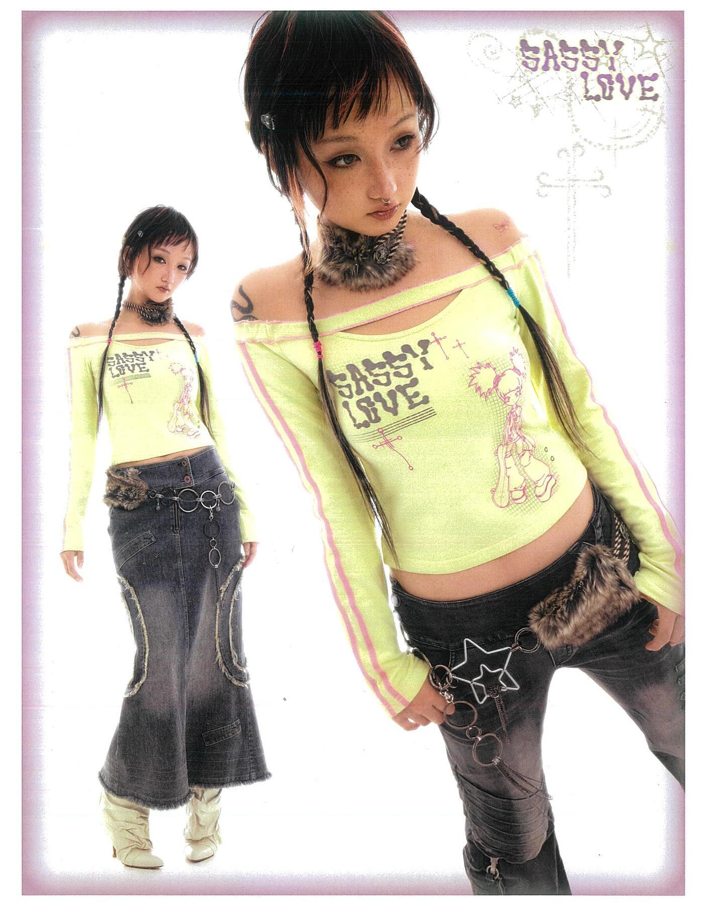 he image showcases two young women, likely models, styled in a distinctly japaneseinspired fashion aesthetic. the dominant style is a blend of harajuku and punk, with pastelcolored clothing and edgy accessories.the primary focus is on a woman wearing a pale yellow, longsleeved, offtheshoulder top with a graphic design that includes the phrase "sassy love."  the top is paired with a dark gray denim skirt featuring an aline silhouette and embellishments. she also wears a dark colored lowrise pants that are darkwash with distressed details. she has long braids and subtle dark eye makeup. the belt she is wearing has a starshaped detail. a similar style is seen on a second girl in the image's background, who is styled similarly but not as prominently.the overall visual is clean, set against a plain white background, enhancing the impact of the clothing and the model's styling. the image possibly comes from a fashion catalog or lookbook.