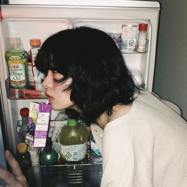 a young woman with short black hair is drinking juice from a carton through a straw while standing in front of an open fridge. she is wearing a white shirt and the fridge is stocked with food and drinks.