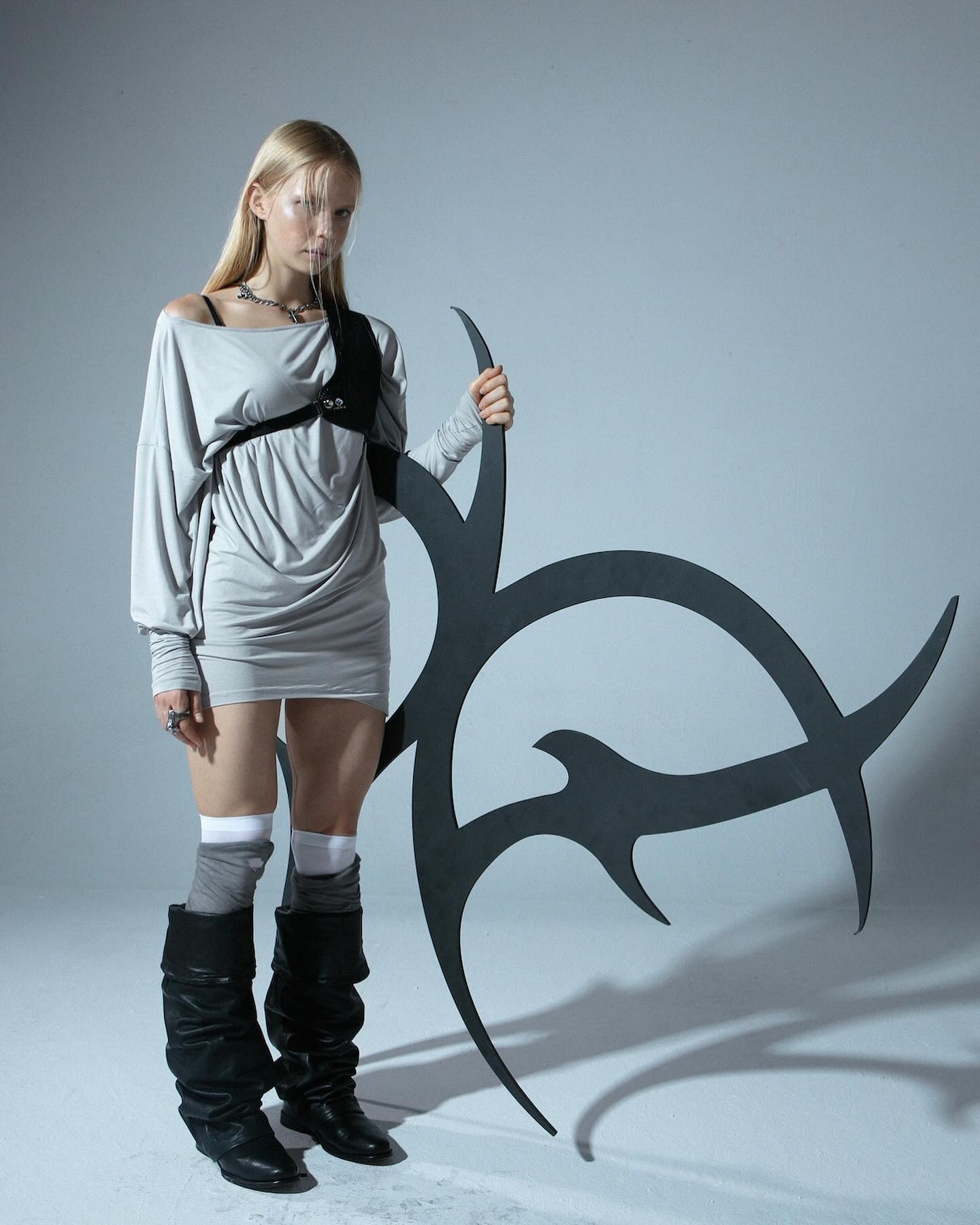 a full shot of a fashion model posing with a large, abstract, dark metal sculpture. she is wearing a loosefitting, light gray longsleeved dress, black slouch boots, and gray socks. she also wears a black shoulder harness and necklace. her blonde hair is straight. the background is a plain light gray studio backdrop. the image has a modern and slightly edgy feel. the model's expression is serious and she appears cool and stylish.