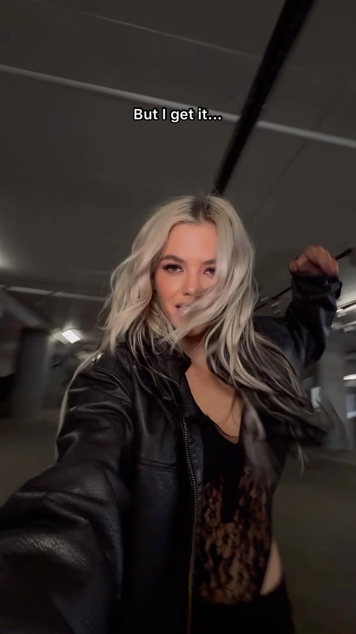 a woman with blonde hair is taking a selfie in an underground parking garage. she is wearing a black leather jacket over a black lace bodysuit. there is text overlaying the image.