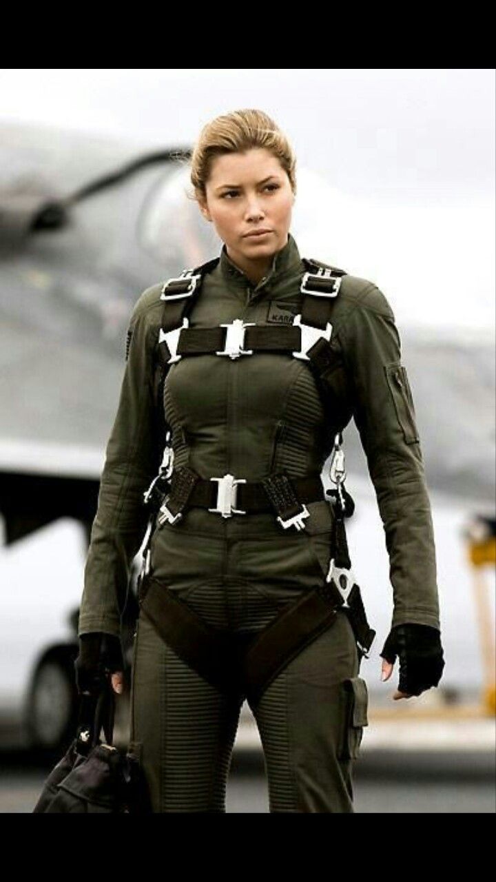 a woman dressed in a military jumpsuit with a harness and gloves, standing in front of a blurry airplane in the background. she appears to be a pilot or a fighter pilot in training.