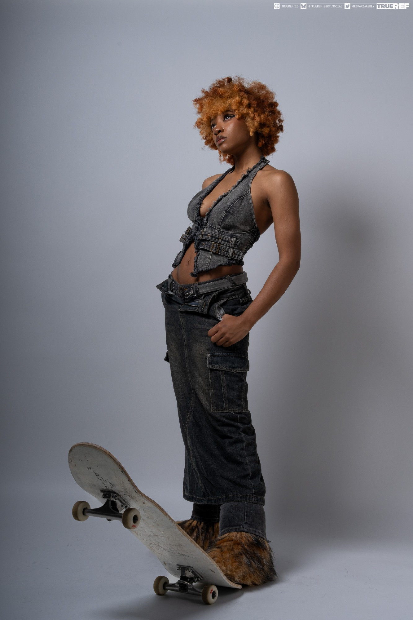 a fullbody shot of a fashion model posing with a skateboard. she is a young woman with curly orange hair, wearing a denim vest, dark cargo pants, and furry boots. she is standing with one leg slightly bent, looking away from the camera with a confident expression.  the backdrop is a simple, neutral grey. the overall style is edgy and urban.