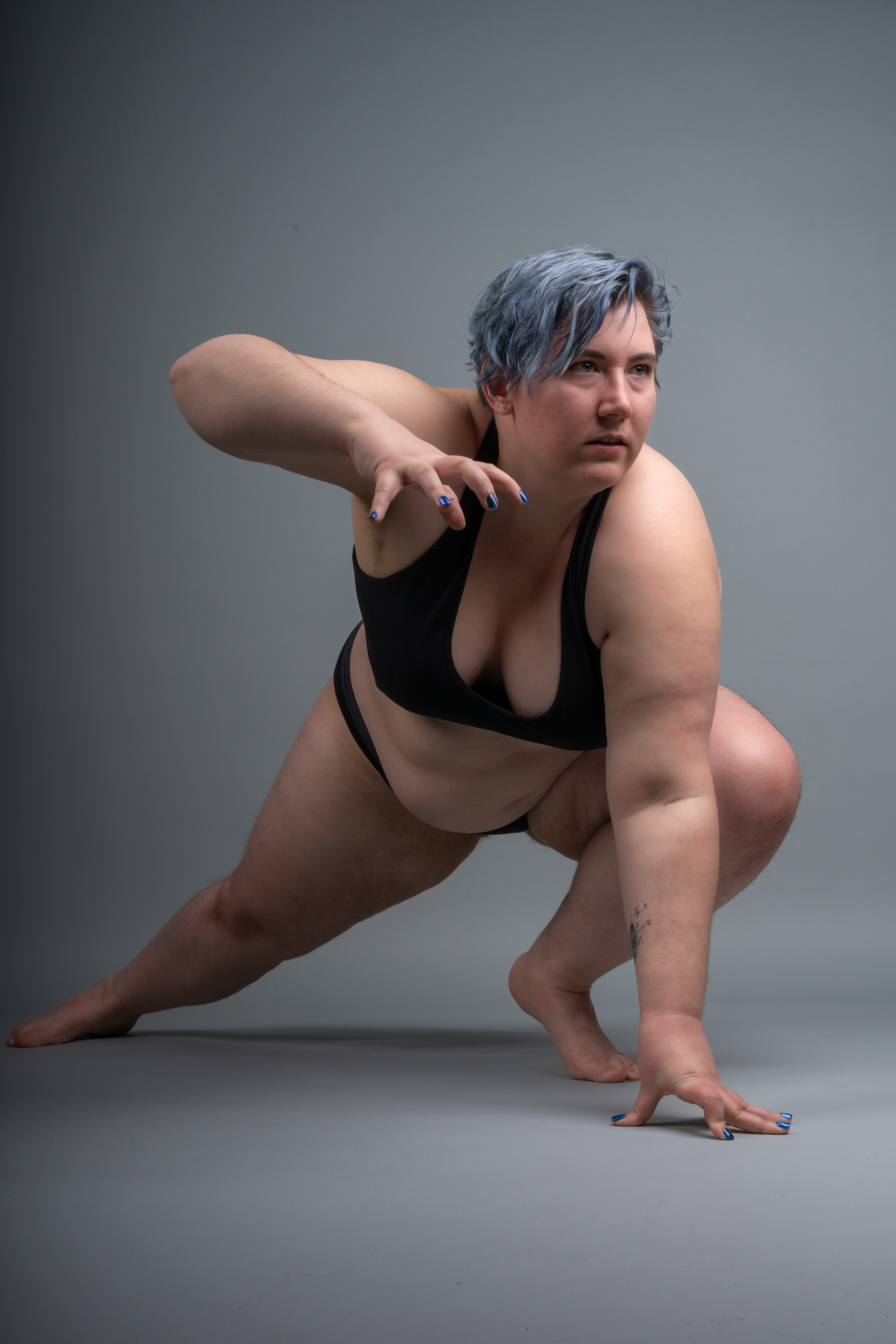 a plus size model is in a crouching pose against a gray background, wearing black lingerie. she is reaching out with one hand, and the other hand is on the ground for support. she has blue hair and is looking away with a confident and fierce expression. there is a tattoo on her arm. it is a studio shot.
