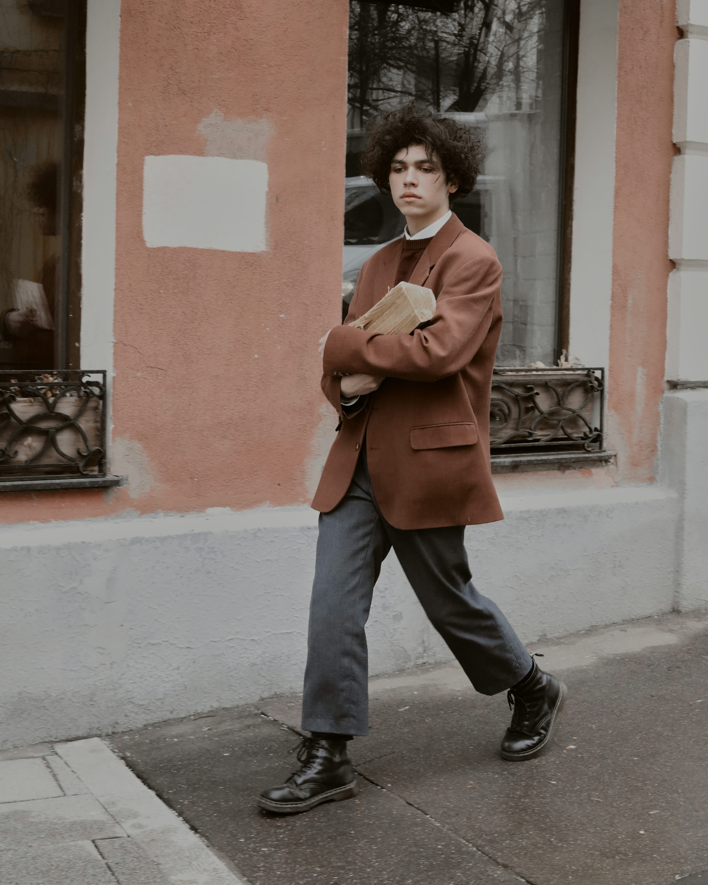 the image shows a young man with curly brown hair wearing a stylish outfit. he's wearing a rustcolored blazer over a white collared shirt, dark gray pants, and black doc martens. he carries a thick book or tome in his arms. the backdrop is an urban setting, with a brick building and a window visible. the overall style and atmosphere of the image are vintage or retro, with a slightly moody and cool feel.  he's walking down a city street and is captured midstride, looking straight ahead.