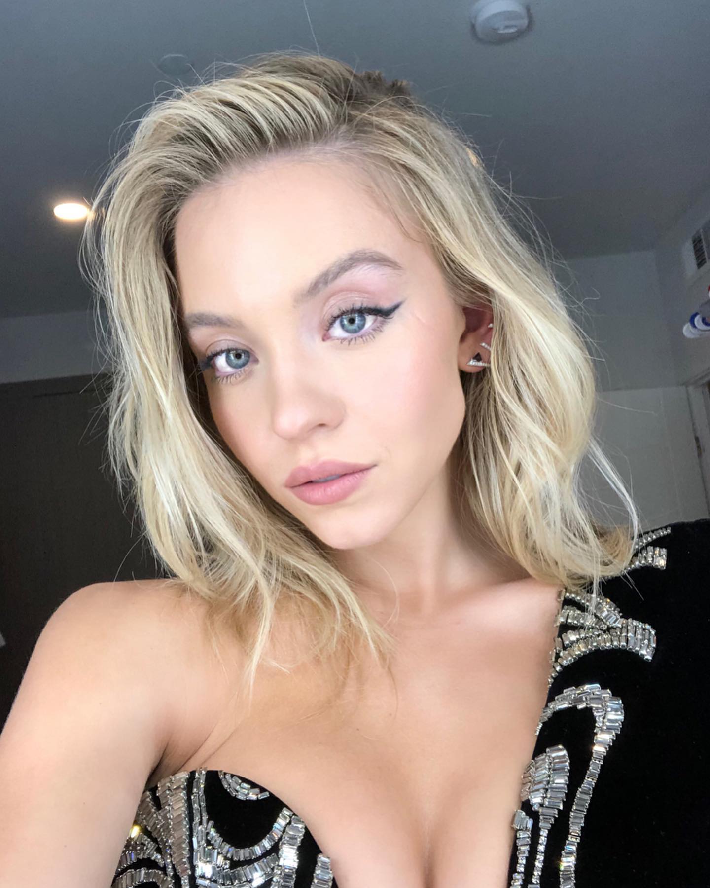 a closeup portrait of actress sydney sweeney. she has blonde, shoulderlength hair styled in waves, blue eyes, and is wearing makeup with winged eyeliner. she is wearing a oneshoulder black dress embellished with silver or white details. the image is taken indoors.