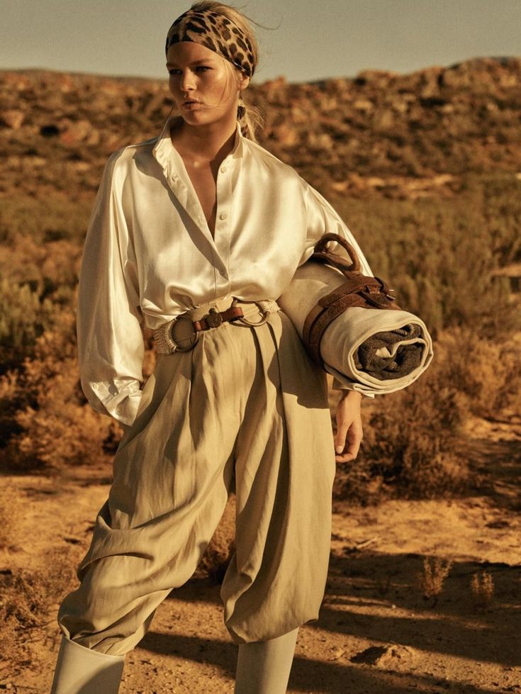 a fashion photograph depicts a female model in a desert landscape. she is wearing a creamcolored silk shirt, highwaisted beige pleated pants that resemble riding breeches, tall white boots, and a leopard print headband.  she carries a rolledup blanket or sleeping bag secured with a brown leather strap. the setting is arid and sandy, with rocky hills in the background. the lighting suggests golden hour, creating a warm and atmospheric image with a vintage or bohochic aesthetic.  the overall style is sophisticated and elegant, fitting for a fashion editorial or campaign.