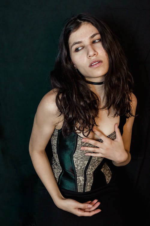 loseup portrait of a young woman with long, dark brown hair. she is wearing a dark green and beige lace corset. her expression is serious and pensive, and she is looking slightly away from the camera. the background is dark and plain. the image has a sensual and elegant feel.