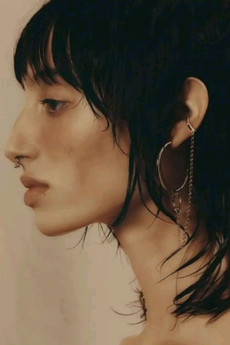 a closeup profile shot of a woman with dark, wet hair and bangs. she has a nose ring and intricate earrings that include a hoop and dangling chain. her bare shoulders are visible, and the overall image has a soft, muted tone.
