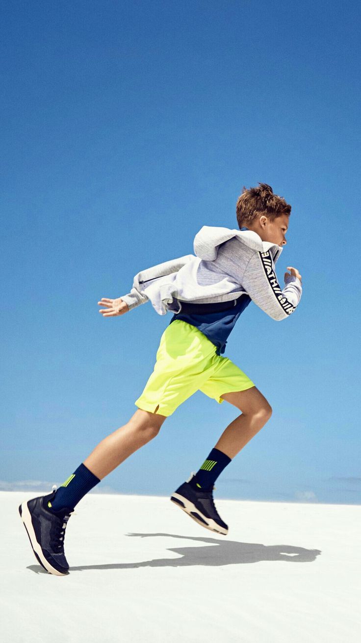 a young boy in grey jacket, neon green shorts, and black sneakers is jumping in the air while running in a blue sky background.  he's wearing black socks with green stripes and a white jacket with black stripes. he is looking toward the right side.  the boy looks like he is having fun while being active.