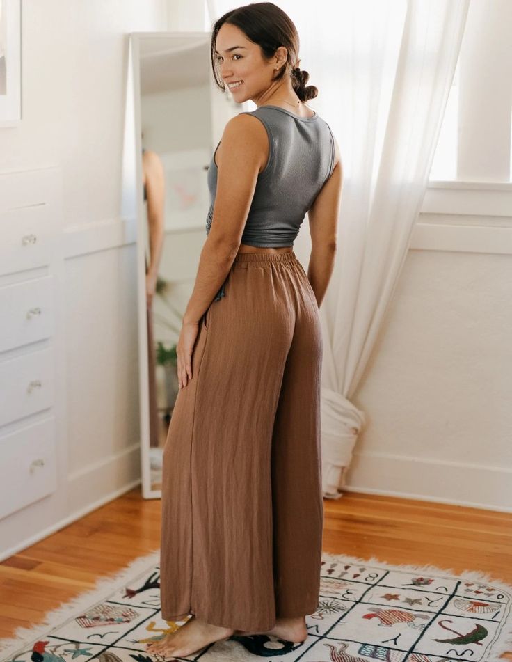a young woman is standing in her bedroom, posing for a mirror selfie. she is wearing a gray crop top and brown wideleg pants. the room is welllit with natural light coming through a window. a colorful rug is laid out on the wooden floor in front of her.