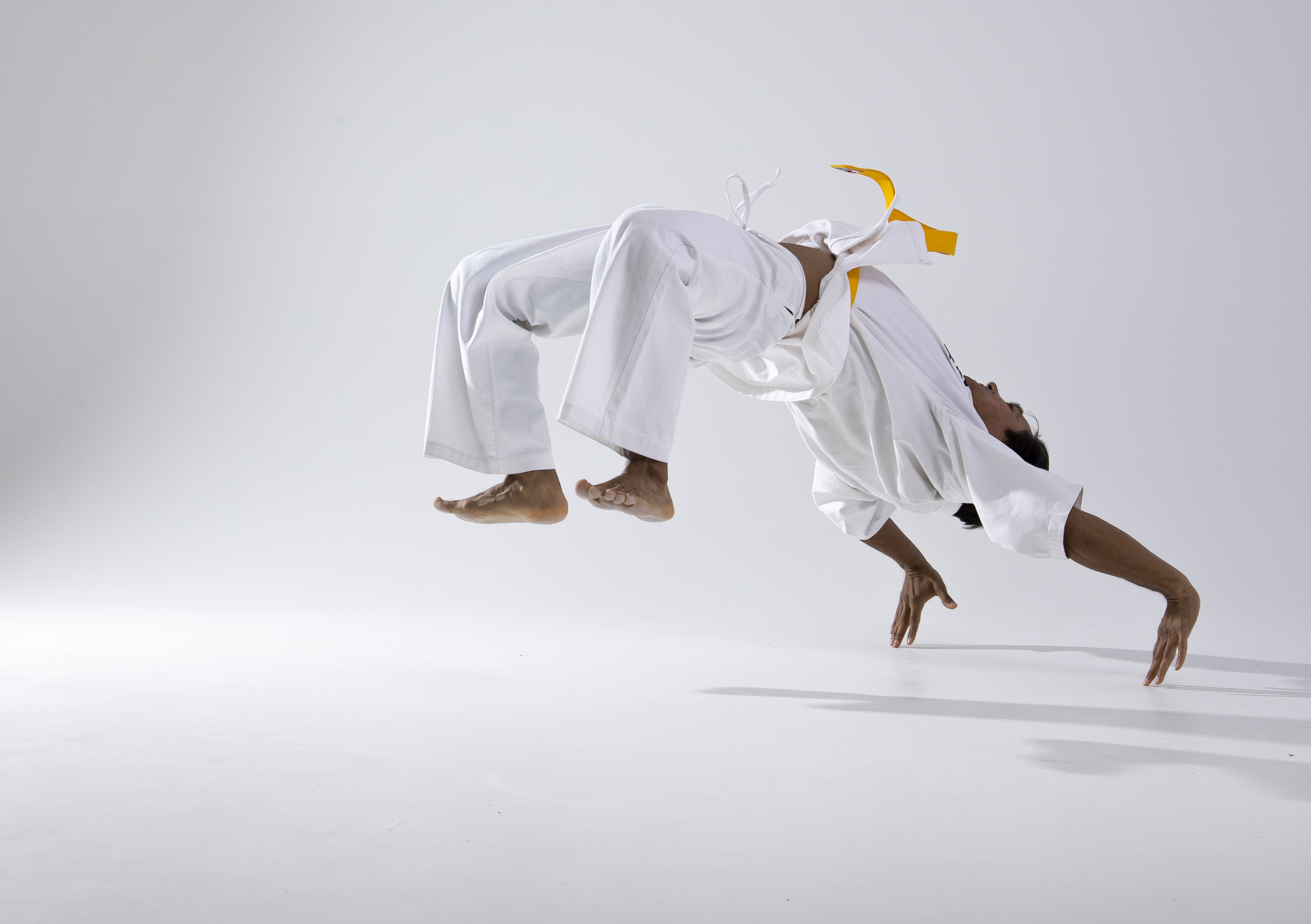 a man in a white uniform with a yellow belt performs a capoeira move in a studio. he is upside down, supported by his hands, with his feet in the air. he is barefoot. the background is white. the scene captures athleticism, agility, and the dynamic movement of capoeira.
