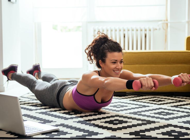 a young woman with curly brown hair tied back in a ponytail is working out in her living room.  she's wearing black and grey workout clothes and a purple sports bra.  laying on her stomach, she is lifting pink dumbbells.  she's smiling at a laptop that's on the floor in front of her.  the laptop is open to a workout video. she's laying on a black and white patterned rug.  behind her is a yellow couch.  the room is well lit with natural light coming from the window behind the couch.