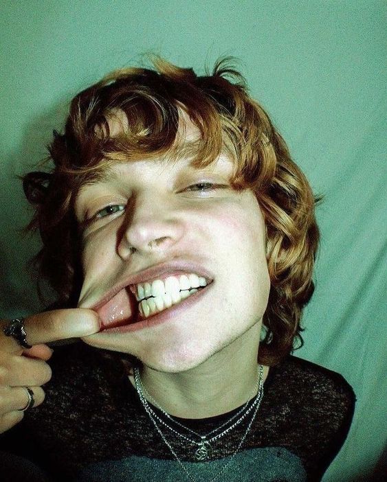 the image is a closeup portrait of a young man with reddish curly hair. he has a nose ring and multiple necklaces. he is pulling his lower lip down with his index finger to reveal his teeth and the inside of his mouth, which is stretched. the lighting is artificial and the background is a teal color.