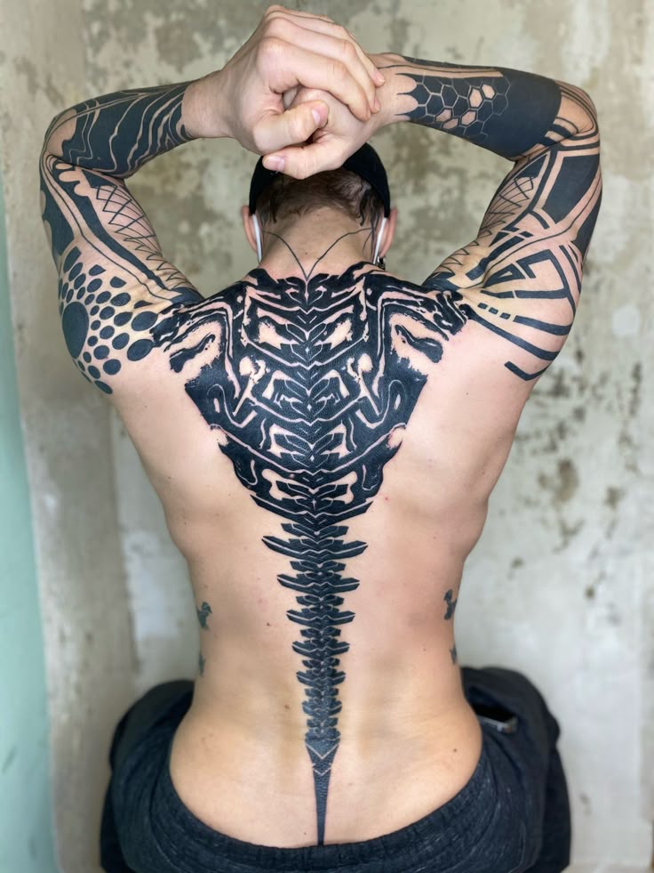 a man is sitting with his arms raised behind his head, showcasing an extensive collection of black ink tattoos. the most prominent tattoo covers his entire back, resembling a stylized spine with intricate geometric patterns. his arms are also heavily tattooed with abstract and geometric designs, indicating a cohesive theme of modern body art. the image captures the detail of the tattoos, highlighting their bold contrast against the man's skin. he is wearing dark casual clothing, and the background appears to be a weathered wall, suggesting an indoor setting.