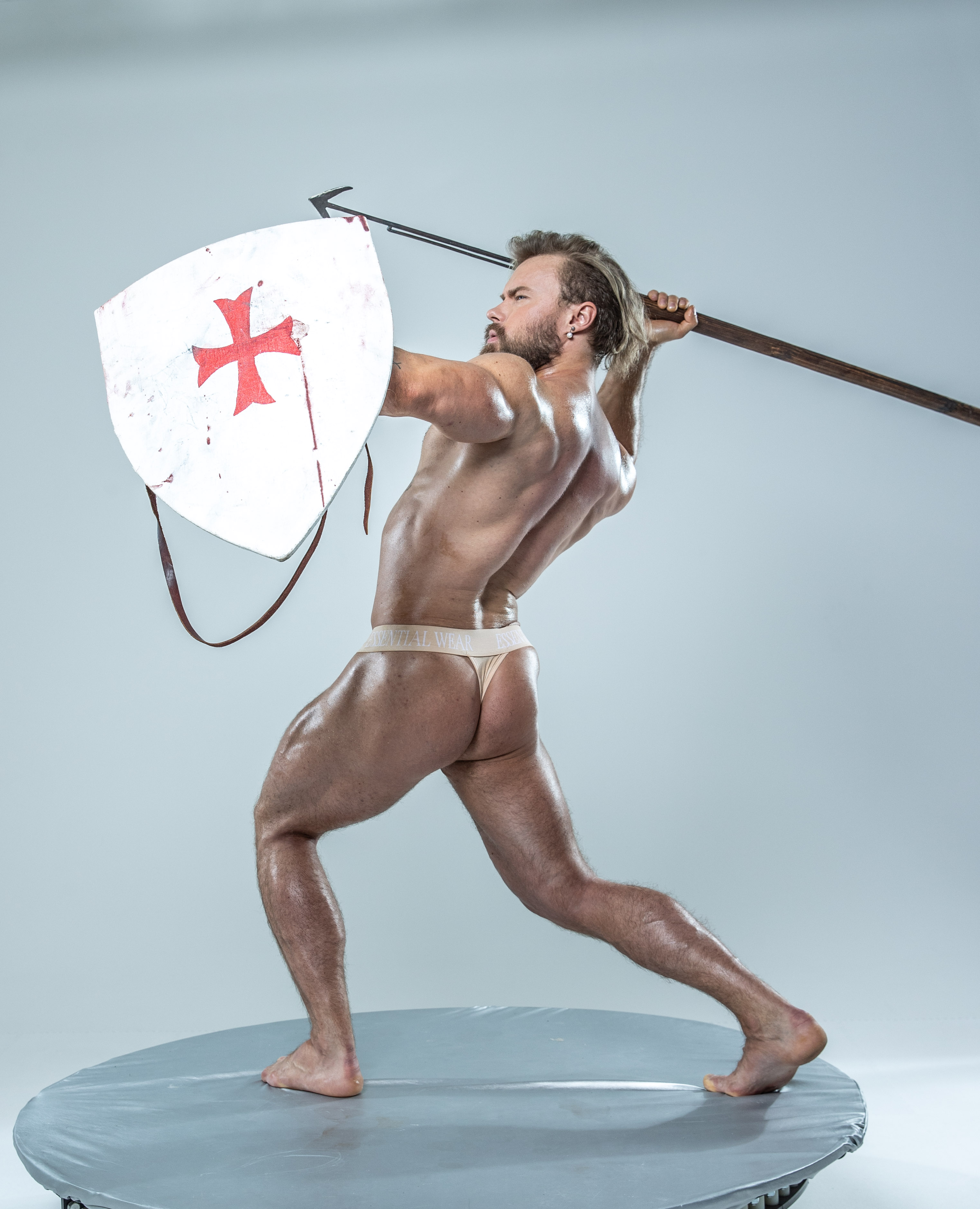 a muscular man, embodying a warrior archetype, stands in a dynamic pose against a light grey backdrop. he wields a spear with a bloodied shield adorned with a red cross, suggesting a historical or mythological context. his powerful physique is emphasized by the studio lighting, showcasing his strength and athleticism.  the low camera angle adds to the image's sense of power and grandeur.