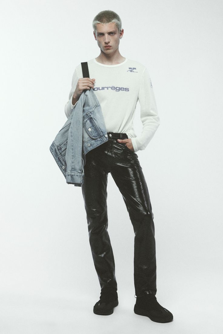 a full shot of a male model standing against a white background. he is wearing a white longsleeved top with the courrèges logo, shiny black pants, and black shoes. he has a denim jacket slung over his shoulder. he is wearing a belt. the shot is taken in a studio.