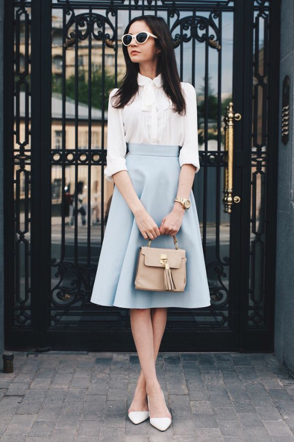 a fullbody shot of a stylish woman standing in front of a black gate. she is wearing a light blue midi skirt, a white blouse with a bow tie, white heels, and white sunglasses. she carries a beige handbag. the setting appears to be outdoors, possibly in a city. the overall style is elegant and chic, suitable for spring or summer.