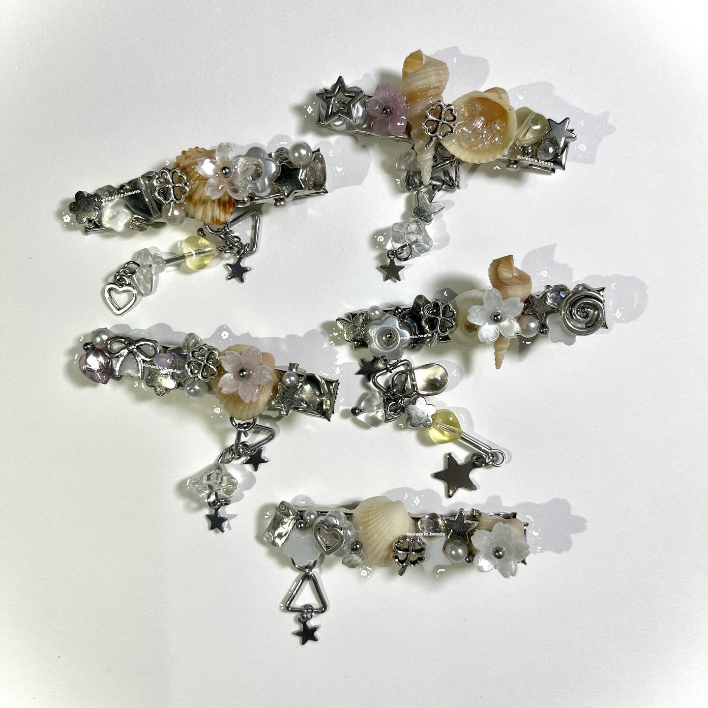 a flat lay of five handmade hair clips decorated with seashells, beads, pearls, flowers, stars, and dangling charms on a white surface. each clip has a unique design.