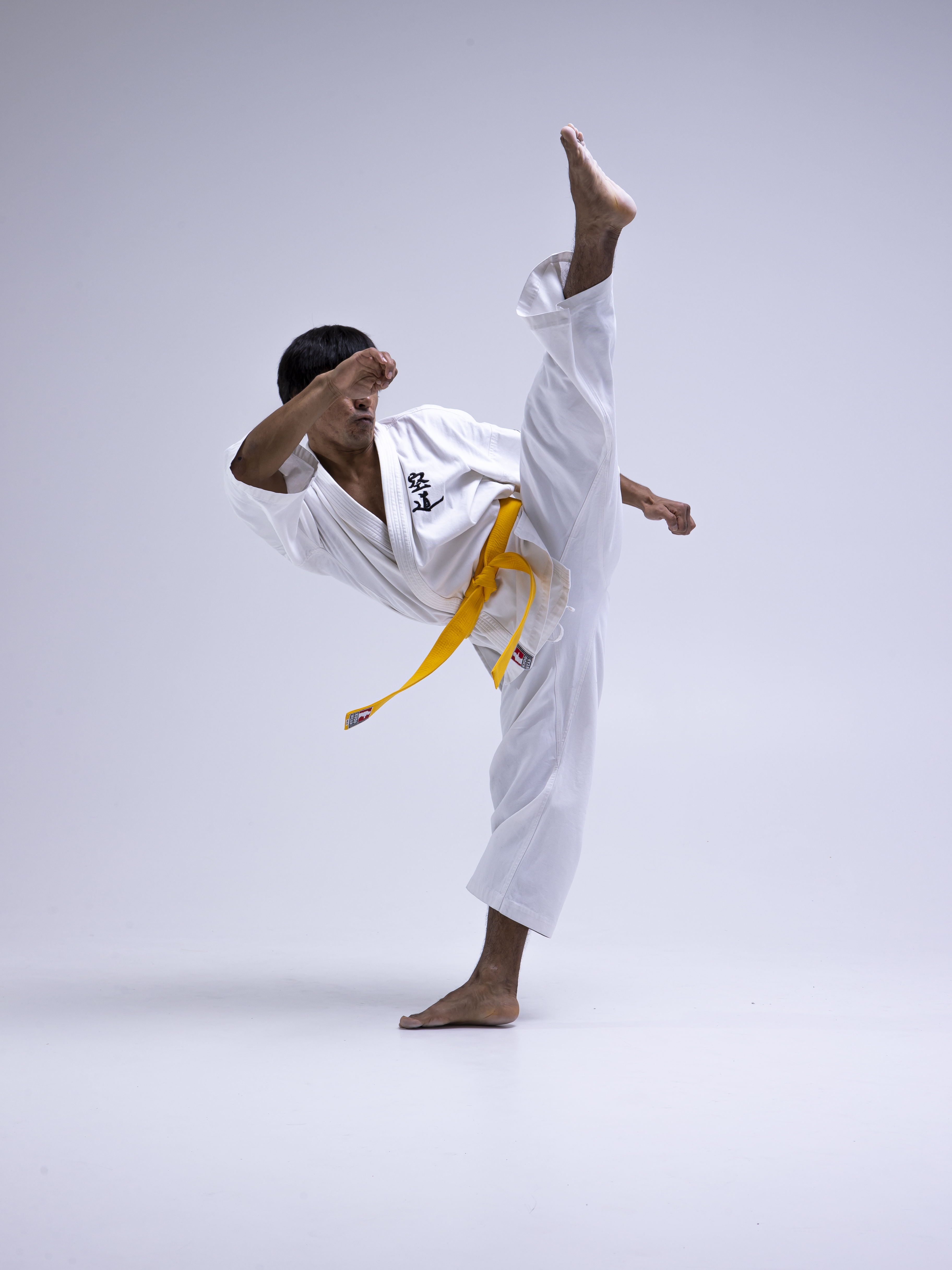 a man is performing a high kick in a karate stance. he is wearing a white karate uniform with a yellow belt. he is barefoot and positioned against a white background in what appears to be a studio setting.