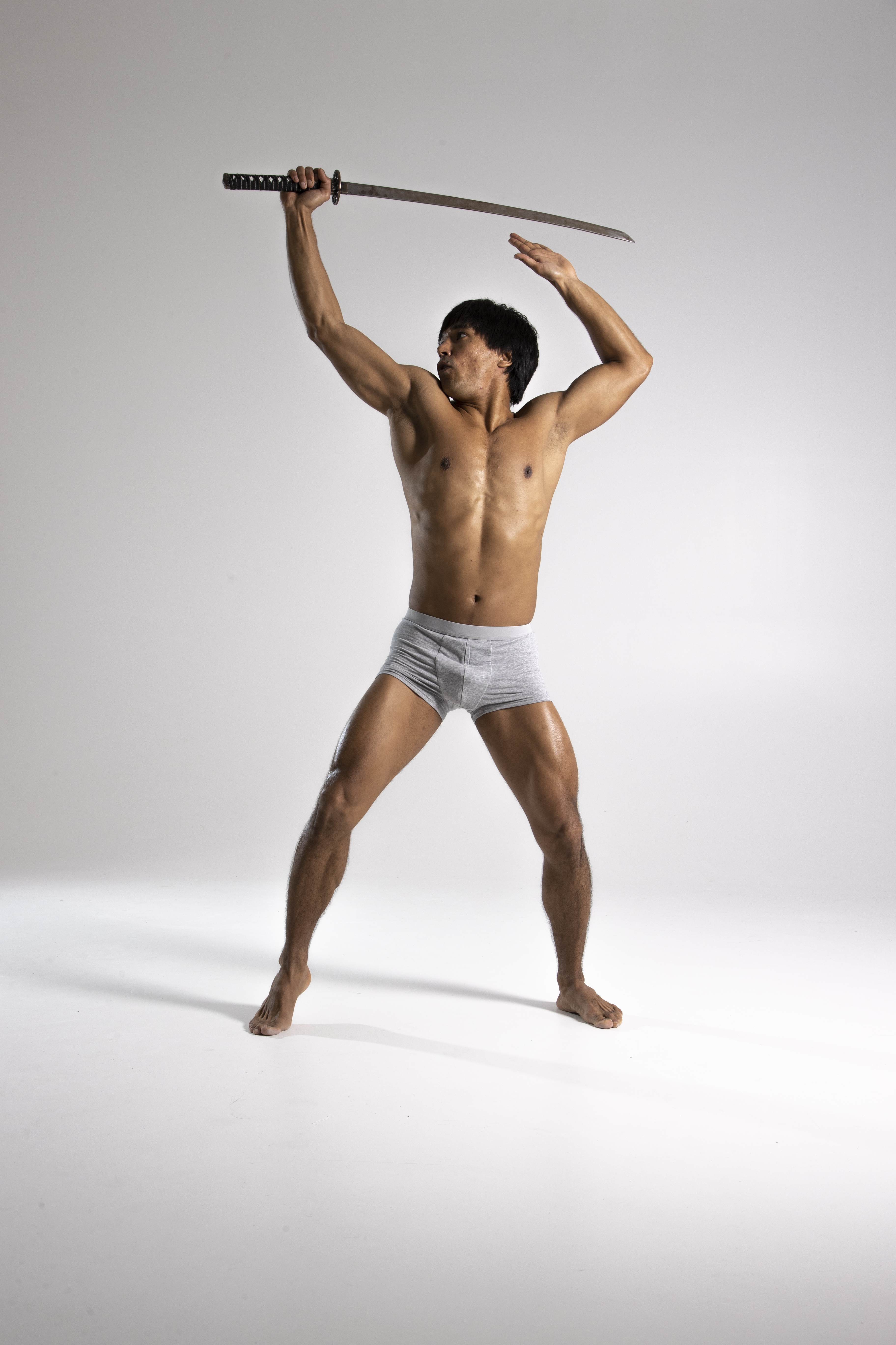 a muscular, barechested man stands against a light gray background, wielding a sword above his head. he is posing, showcasing his athletic build. he is wearing light gray underwear.