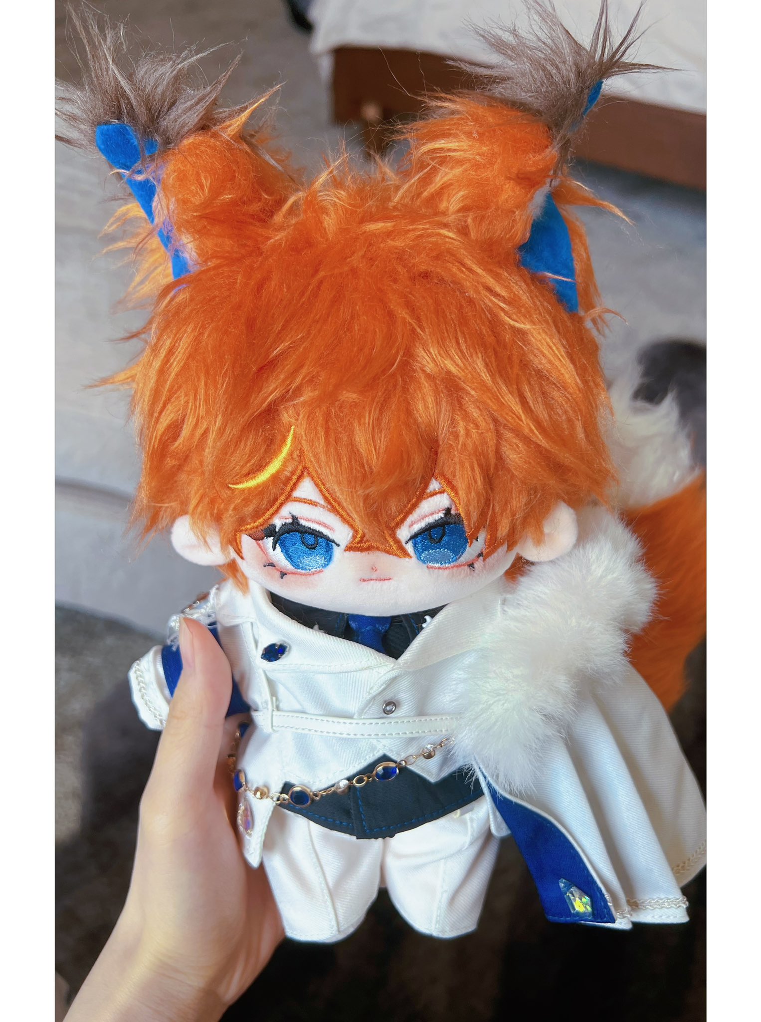 a plush toy anime character with orange fur hair, blue eyes, a white coat with a blue ribbon, is being held in a person's hand.